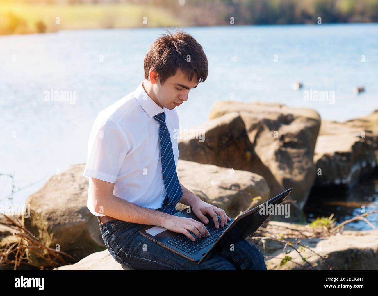 Un garçon portant une chemise blanche, une cravate et un Jean  étudiant/apprenant en ligne sur l'ordinateur à l'extérieur près du lac. Vue  de l'arrière Photo Stock - Alamy