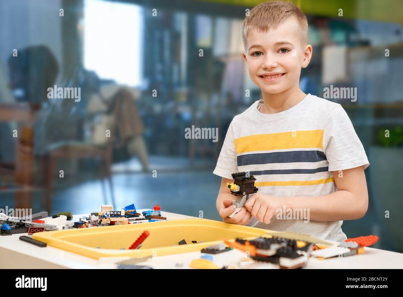 Vue de face d'un charmant garçon caucasien souriant et regardant l'appareil photo. Kit de construction pour les enfants sur la table, les enfants créant des jouets, ayant des émotions positives et Banque D'Images