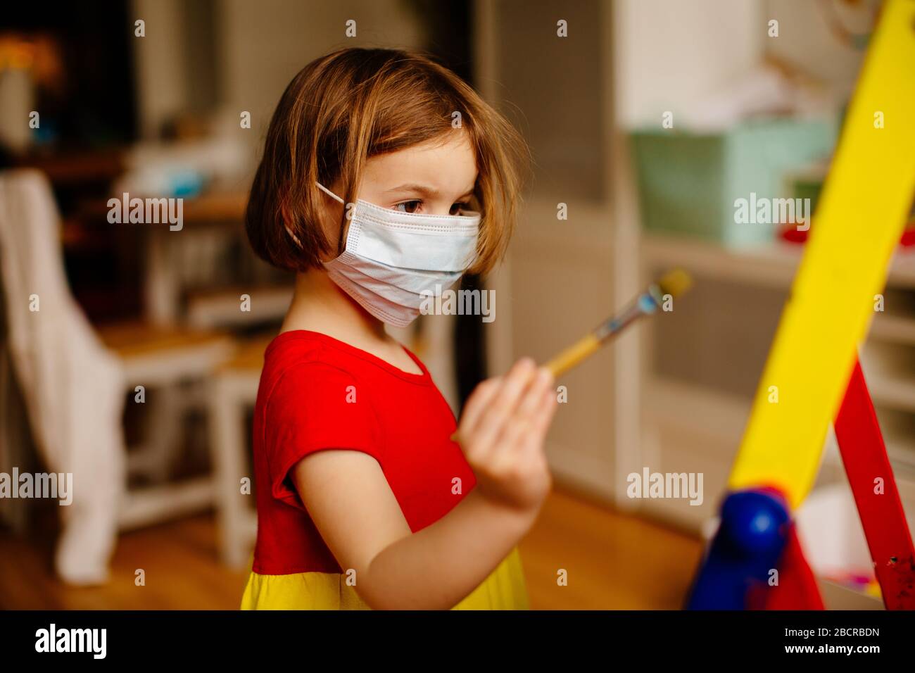 Portrait d'un petit enfant préscolaire avec protection antivirus masque chirurgical du visage en raison du coronavirus, peinture à un chevalet d'art à la maison Banque D'Images