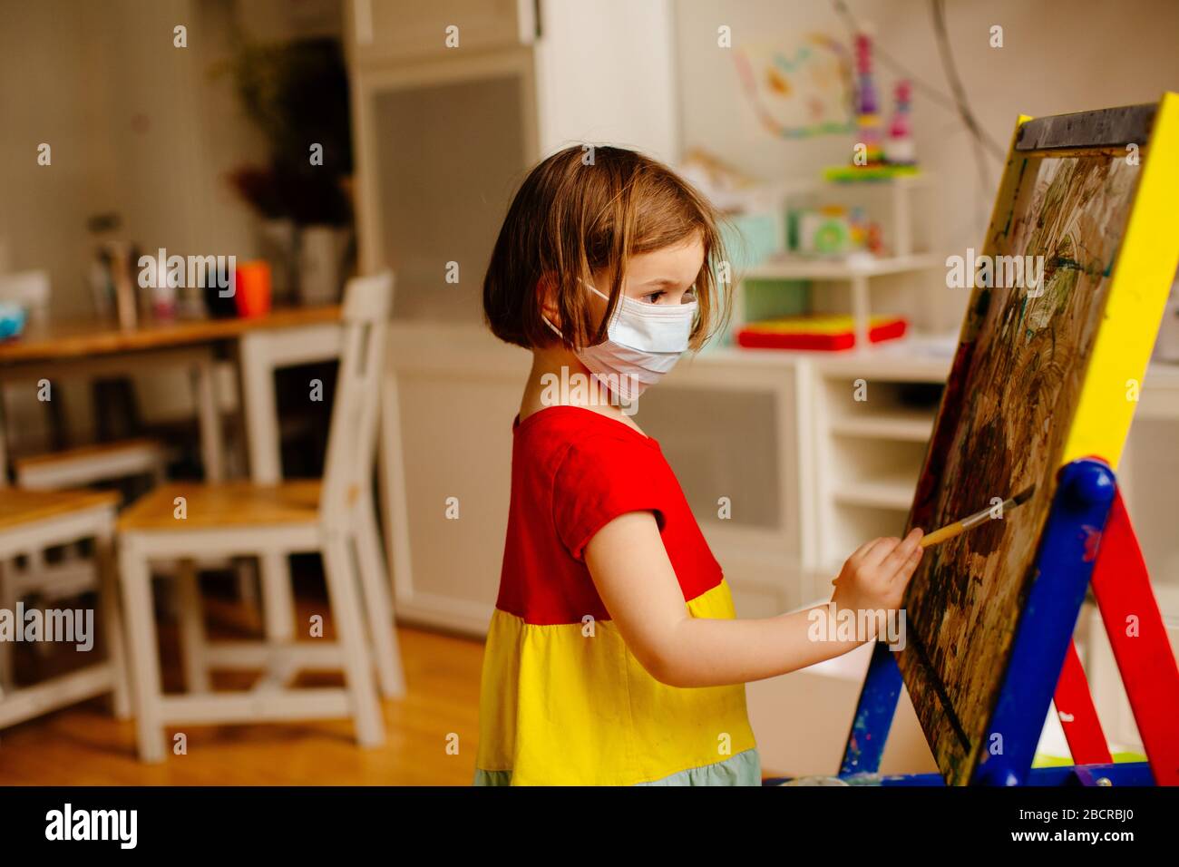 Portrait d'un petit enfant préscolaire avec protection antivirus masque chirurgical du visage en raison du coronavirus, peinture à un chevalet d'art à la maison Banque D'Images