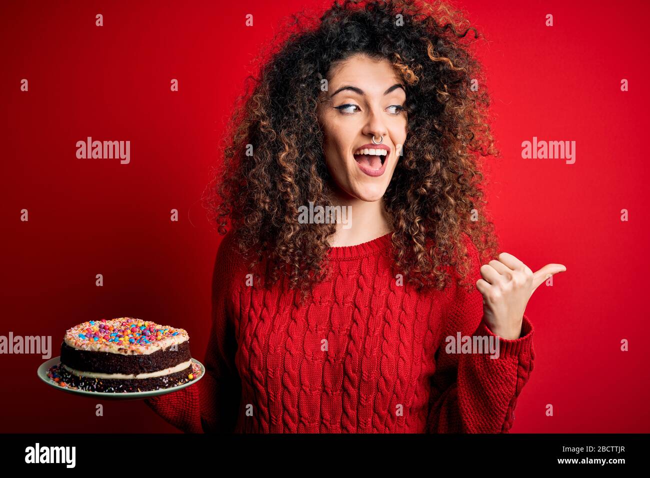 Jeune Femme Belle Avec Des Cheveux Boucles Et Une Plaque De Conservation Piercing Avec Gateau D Anniversaire Pointant Et Montrant Avec Le Pouce Jusqu Au Cote Avec Le Visage Heureux Smi Photo Stock