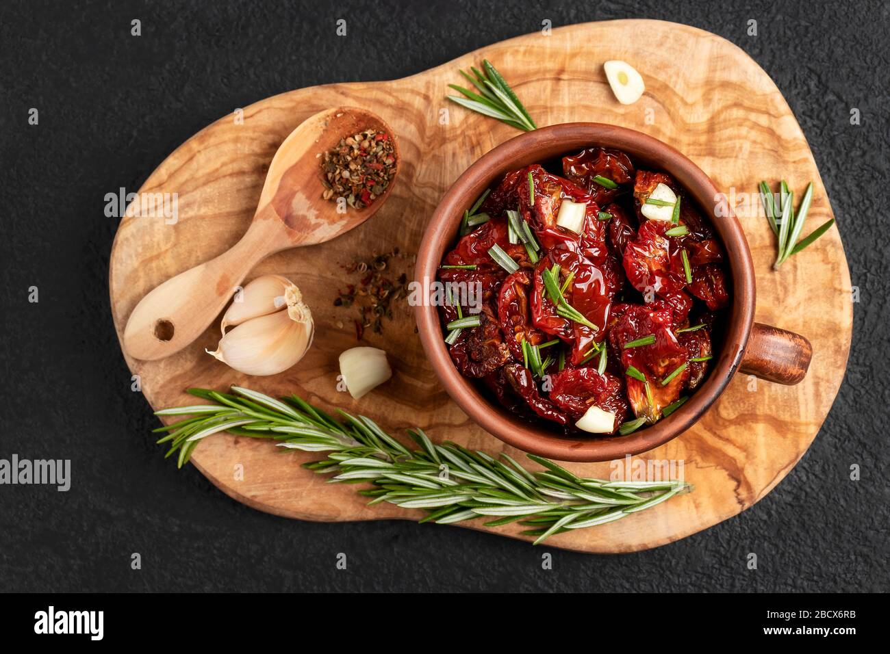 Tomates séchées au soleil avec ail, romarin et épices dans un bol en argile sur un tableau de découpe en bois d'olive, vue sur le dessus, plat. Banque D'Images