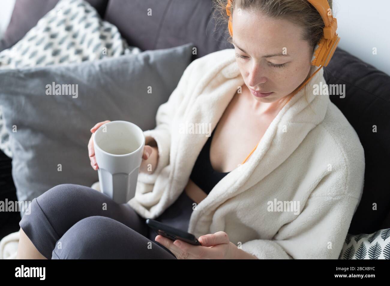 Restez à la maison. Distanciation sociale. Femme à la maison se reposant sur un canapé canapé canapé-lit boire du thé de la tasse blanche, écouter de la musique relaxante, rester connecté à Banque D'Images