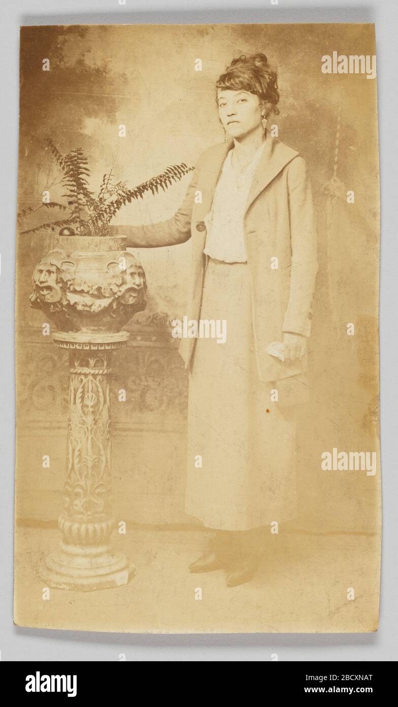Carte postale photographique d'une femme. Une carte postale photographique d'une femme debout avec sa main au sommet d'un vase. La photo pleine longueur de la femme présente sa position devant un fond peint portant une jupe longue et une veste longue et des boucles d'oreilles. Carte postale photographique d'une femme Banque D'Images