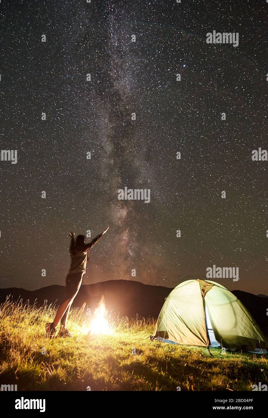 Happy femme randonneur se reposer au camping de nuit d'été à côté d'un feu de camp et de la tente touristique laverante. Jeune femme tenant les mains se lever, profitant de la vue du ciel de nuit plein d'étoiles et de voie lactée. Banque D'Images