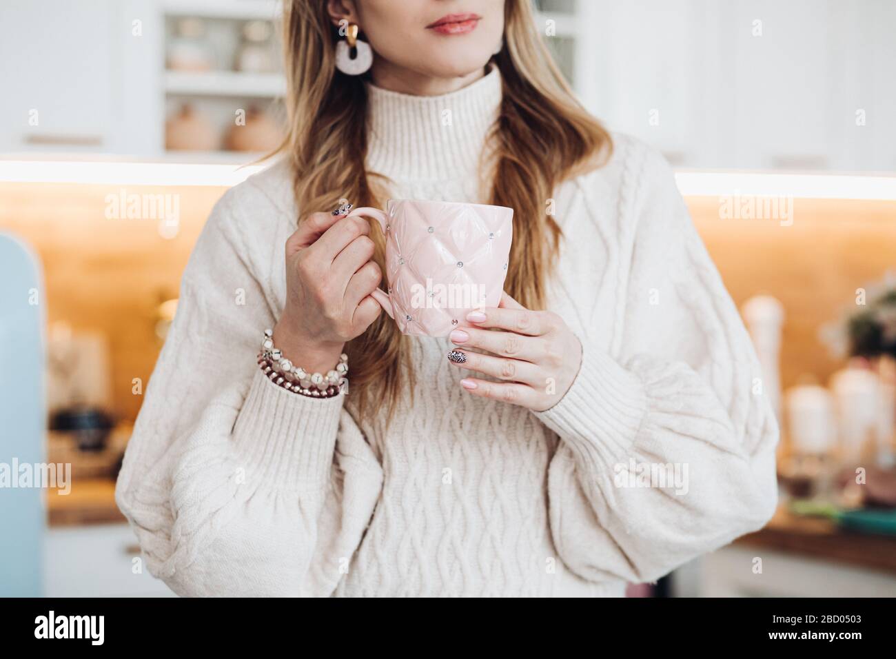 Photo courte d'une jeune femme élégante dans un pull confortable et des bracelets tenant un joli mug rose avec des strass dans ses mains soignées dans le Banque D'Images