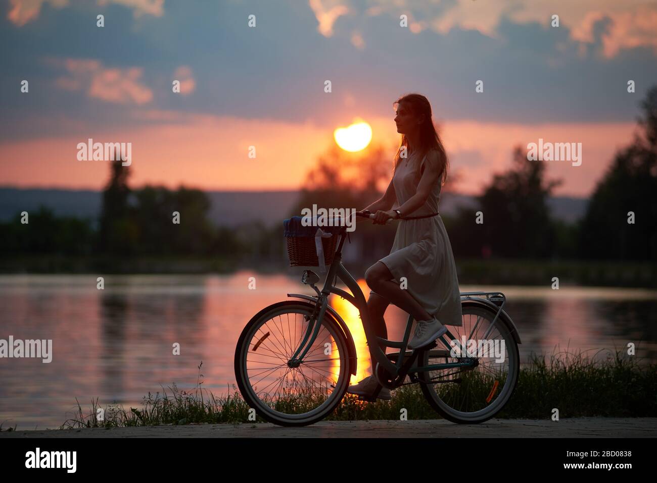 Jolie femme en robe conduit un vélo vintage avec panier sur la route près de la rivière au coucher du soleil ciel avec des nuages. Soirée d'été jour au coucher du soleil rose et bleu ciel arrière-plan en plein air Banque D'Images