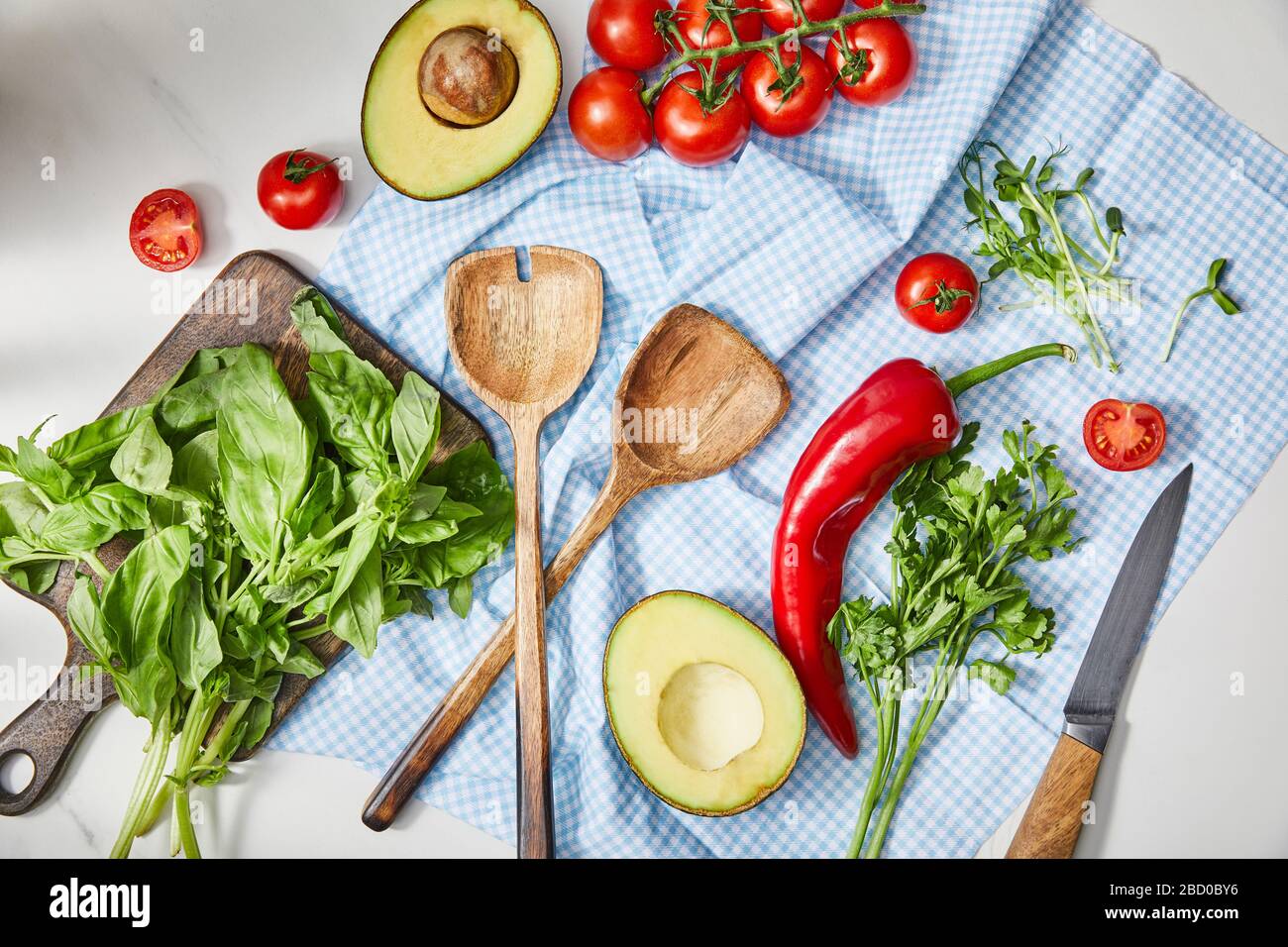 Vue de dessus des légumes, de la verdure, des moitiés d'avocat, des couteaux et des spatules sur le tissu près du basilic sur la planche à découper en blanc Banque D'Images