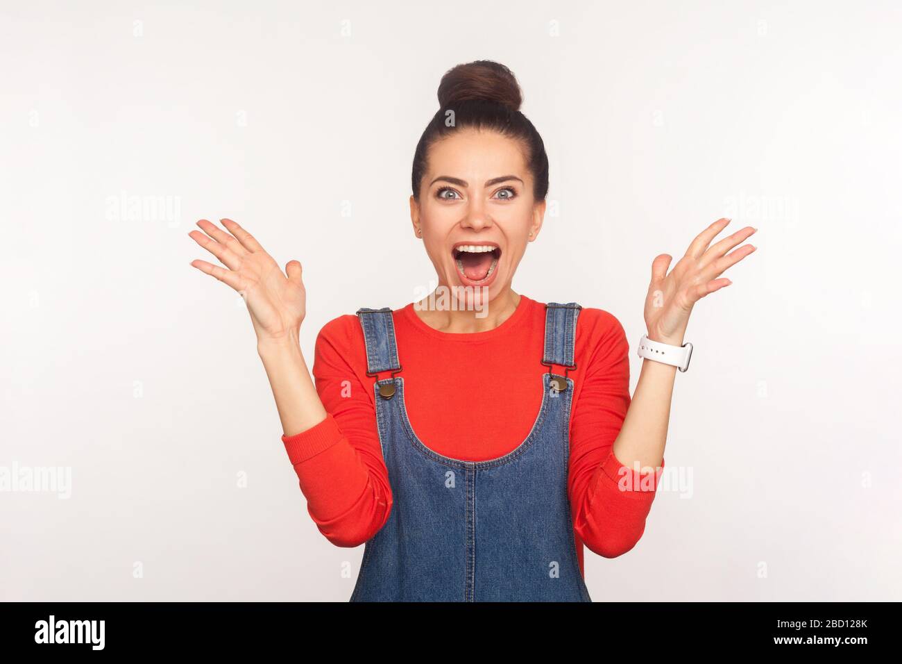 OH mon dieu, wow! Portrait d'une jolie fille joyeuse surprise avec un pain de cheveux dans des salopettes en denim qui a l'air impressionné par des nouvelles incroyables, levant les mains Banque D'Images