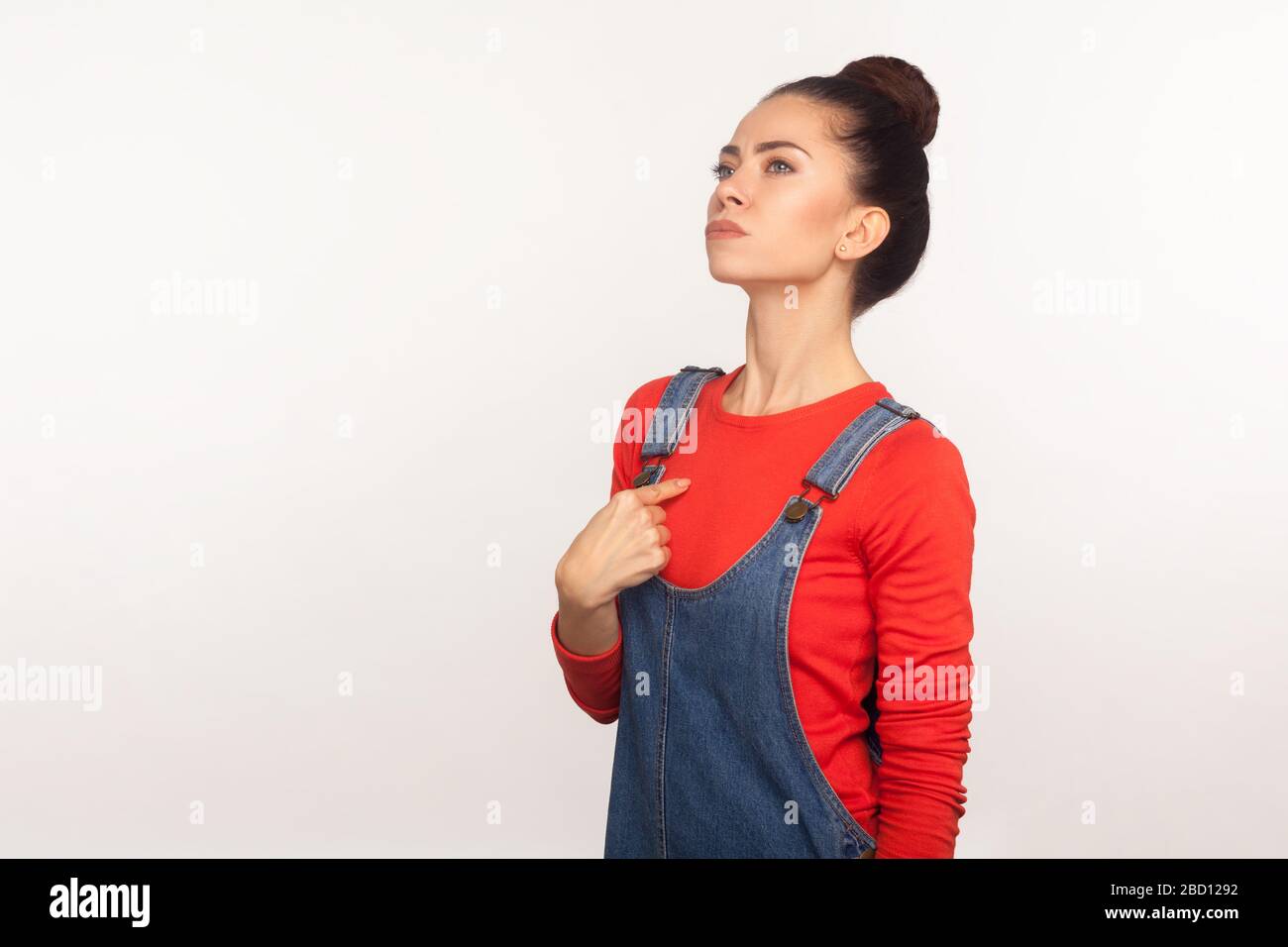 Portrait d'une fille arrogante égoïste avec un pain de cheveux dans des salopettes en denim pointant vers elle et regardant avec fierté l'expression de soi-même, exagérée Banque D'Images