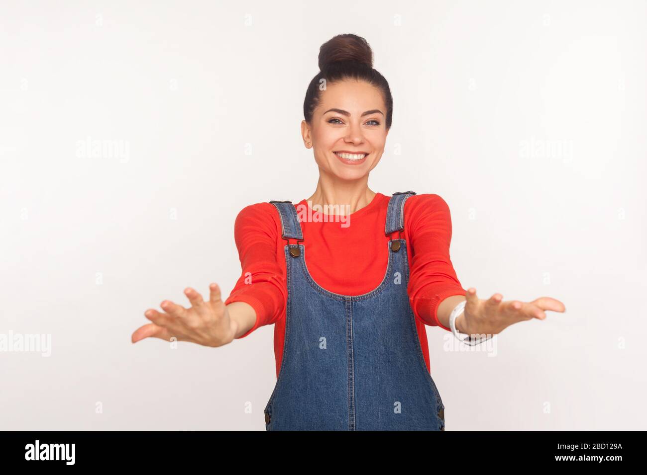 S'il vous plaît, prenez-le! Portrait d'une fille positive et élégante avec un pain à cheveux dans des salopettes en denim invitant à embrasser, garder les mains comme donner, partager gratuitement. ind Banque D'Images