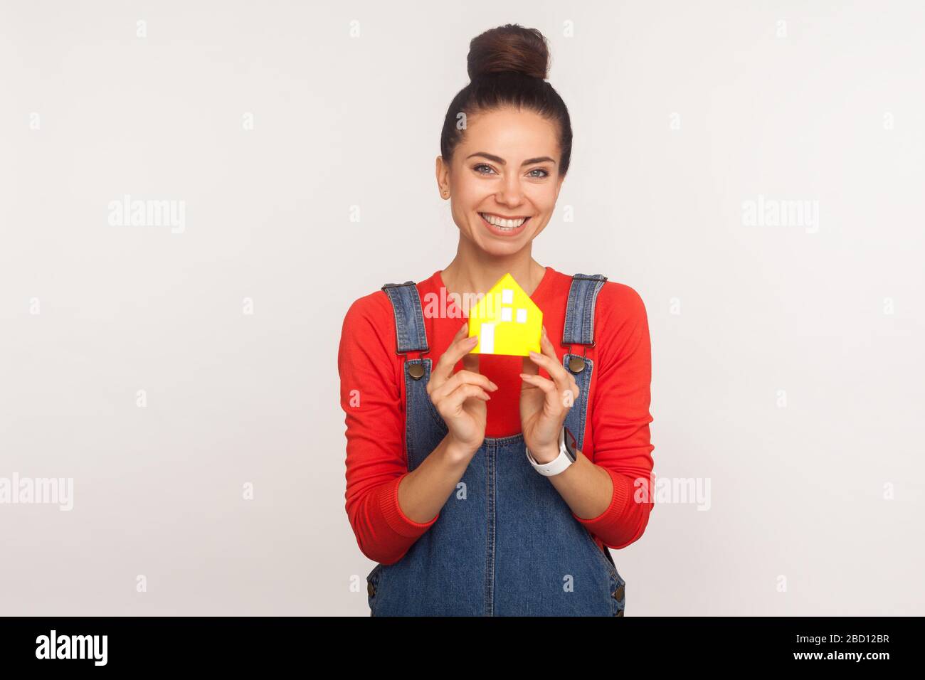 Achat à domicile. Portrait d'une charmante fille optimiste avec un pain à cheveux dans des combinaisons en denim de style urbain tenant maison en papier et regardant l'appareil photo avec toothy Banque D'Images