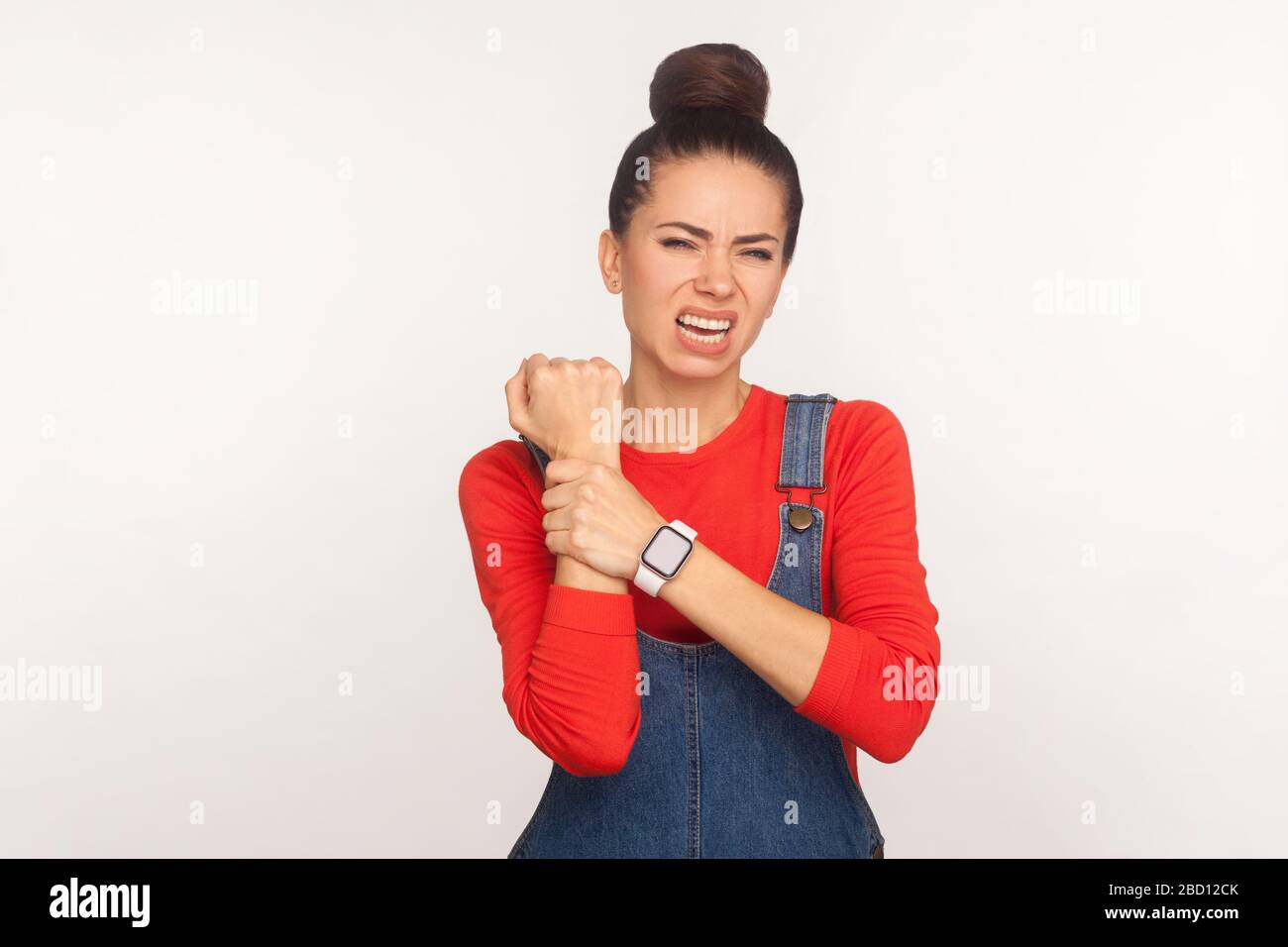 Poignet tentaculaire, rigidité musculaire. Portrait d'une fille malsaine avec un pain de cheveux dans des salopettes en denim massant la main douloureuse, souffrant du syndrome du tunnel carpien. Banque D'Images