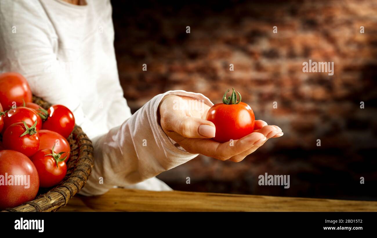 Main de femme tenant la tomate rouge et le fond de table en bois avec des tomates mûres fraîches dans un bol de panier en osier et un espace de copie pour la nourriture et les produits. Banque D'Images