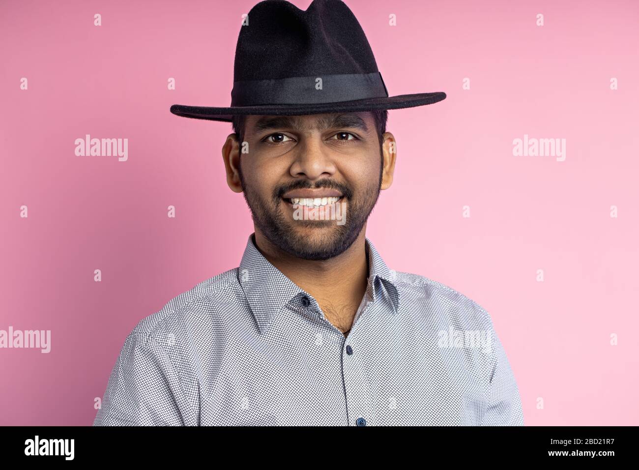 Gros plan de jeune homme indien joyeux et heureux avec barbe de 3 jours, portant une chemise décontractée, un chapeau noir, regardant avec le sourire et pointant sur le côté avec l'index Banque D'Images