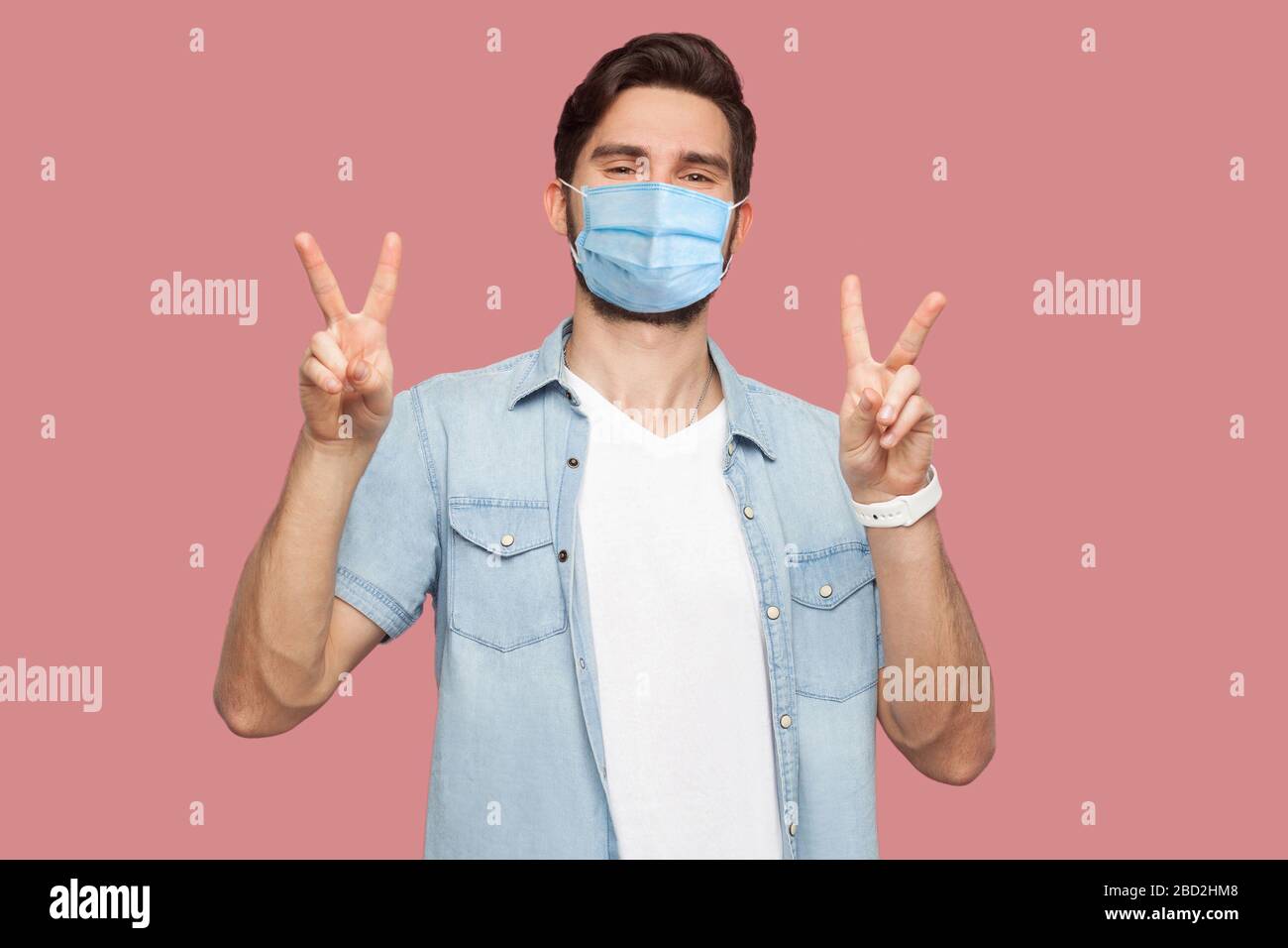 Portrait d'un jeune homme heureux avec masque médical chirurgical en chemise bleue debout avec victoire ou signe de paix main geste et regardant l'appareil photo avec le sourire Banque D'Images