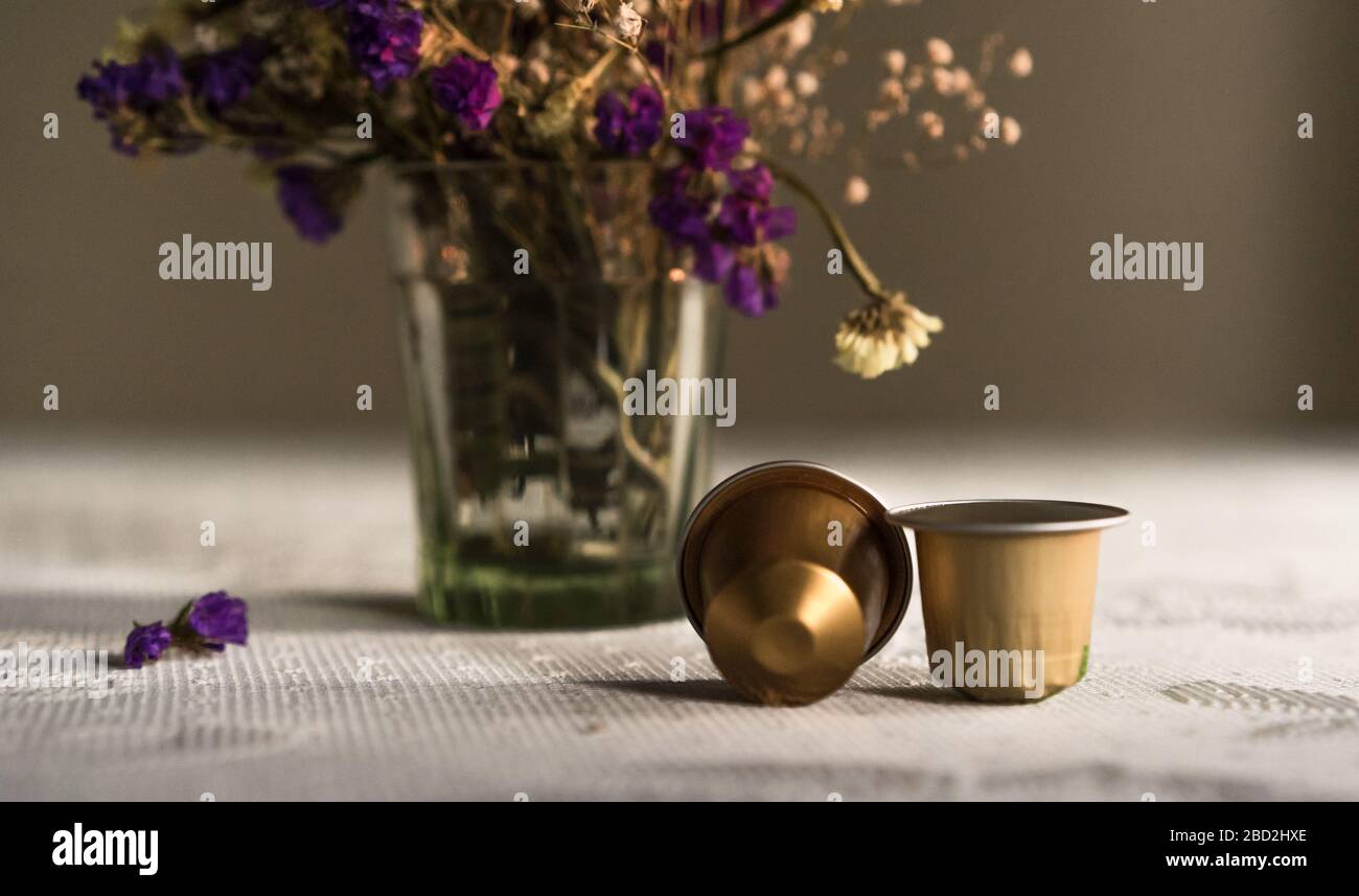 Deux capsules de café Nespresso sur la table devant les fleurs violettes et blanches dans un vase en verre textile blanc sur fond sombre Banque D'Images