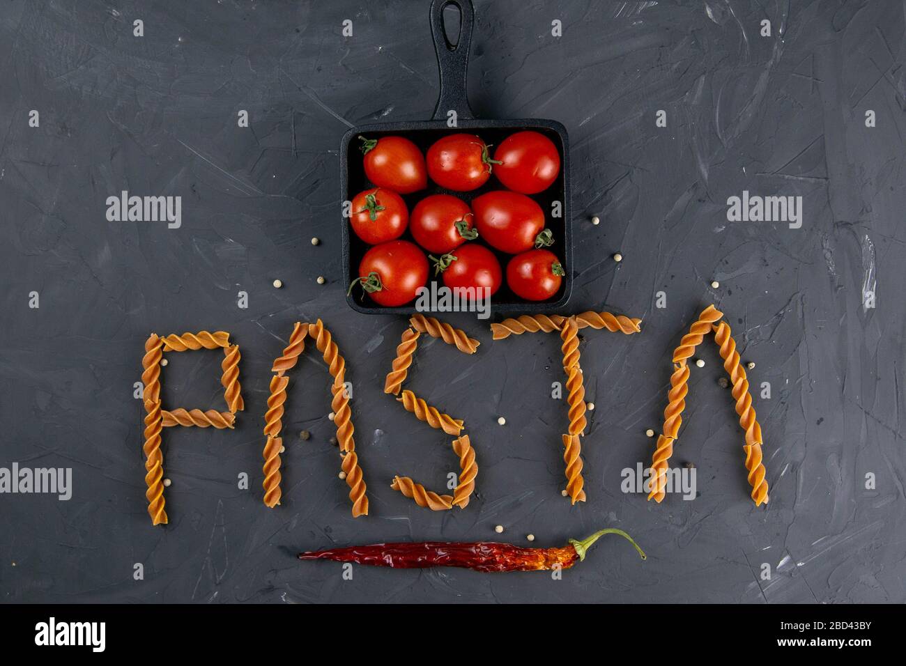 Fusilli italien - mot pâtes. Pâtes italiennes crues fusilli avec tomates cerises dans la poêle à frire et papier chaud sur fond sombre d'en haut. Pose plate Banque D'Images