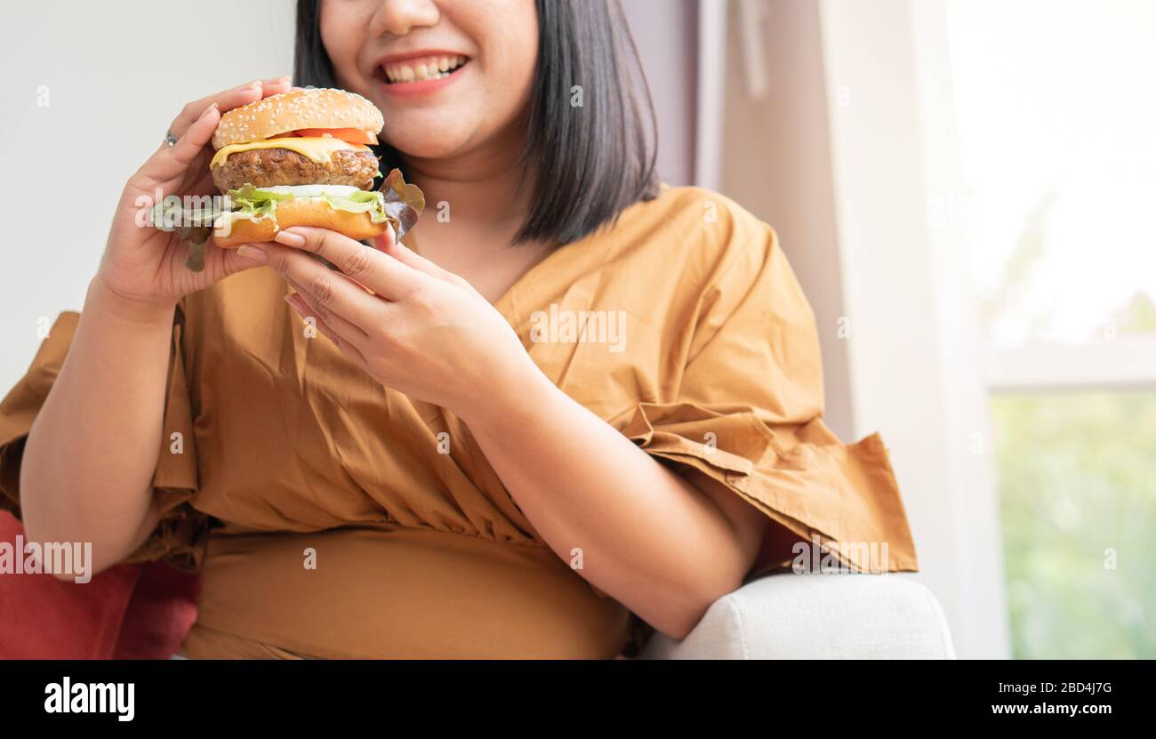 Faim surpoids woman smiling and holding hamburger et assis dans le salon, son très heureux et profiter de manger les aliments de préparation rapide. Concept de binge eati Banque D'Images