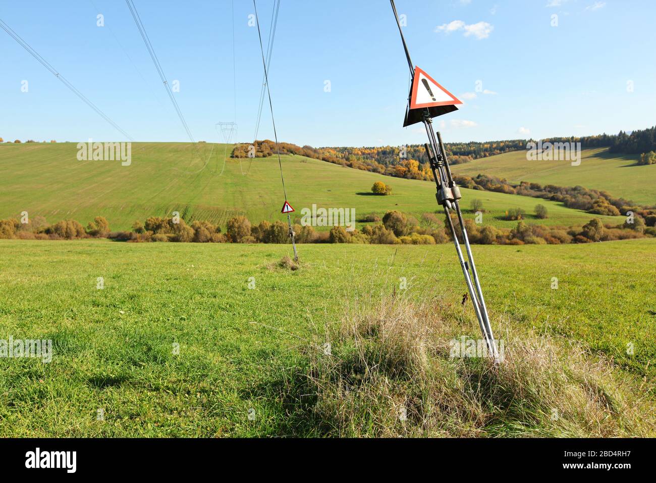 Câbles en acier au pylon électrique avec triangles d'avertissement rouges avec point d'exclamation - danger, haute tension, petites collines avec prés verts Banque D'Images