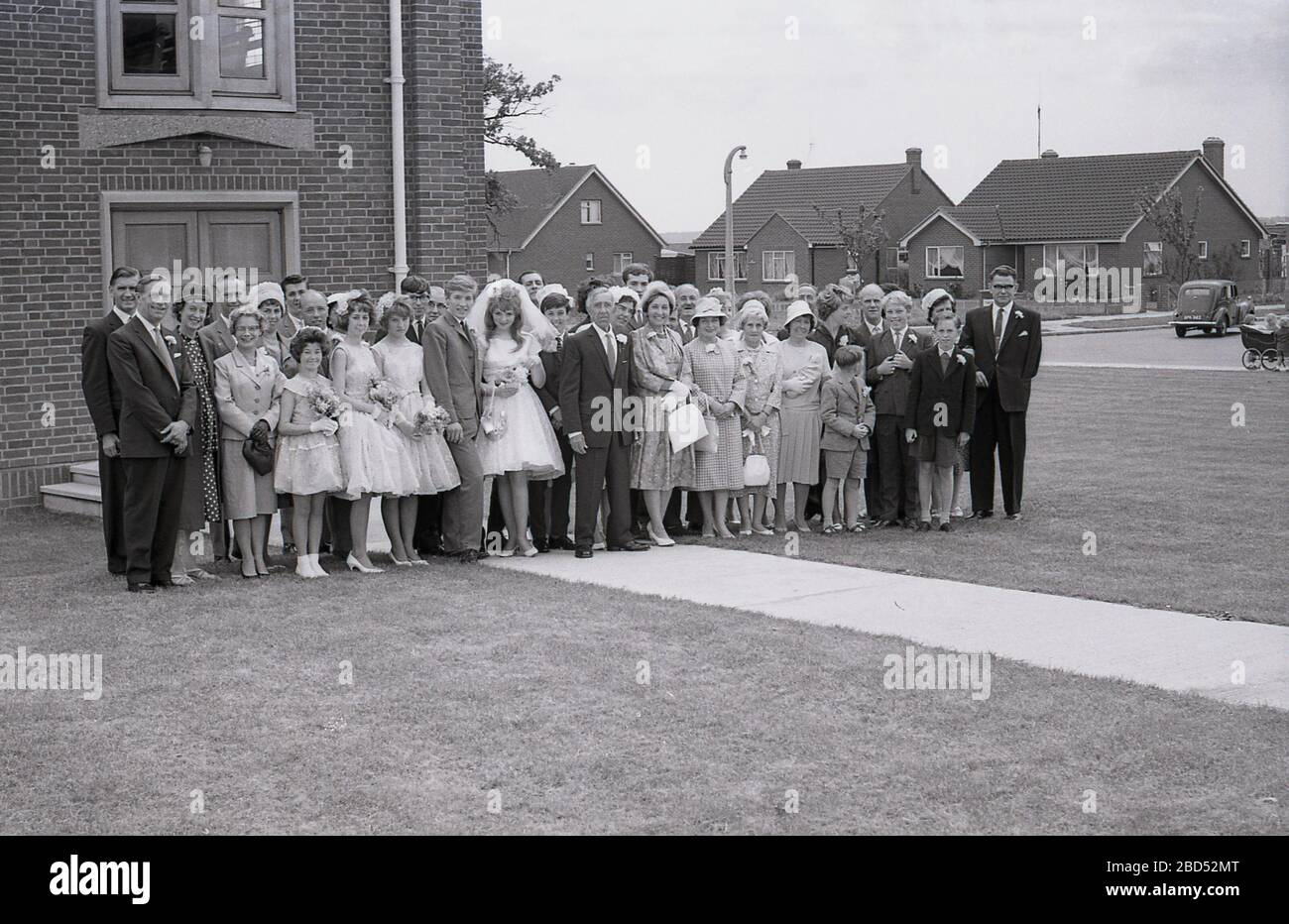 Années 60, historique, sur un immobilier de banlieue, à l'extérieur d'une église moderne, une photo de groupe d'une mariée et d'une marié d'adolescence et de leurs invités de mariage, avec une voiture de voiture et un landau de l'époque à distance, Angleterre, Royaume-Uni. Banque D'Images