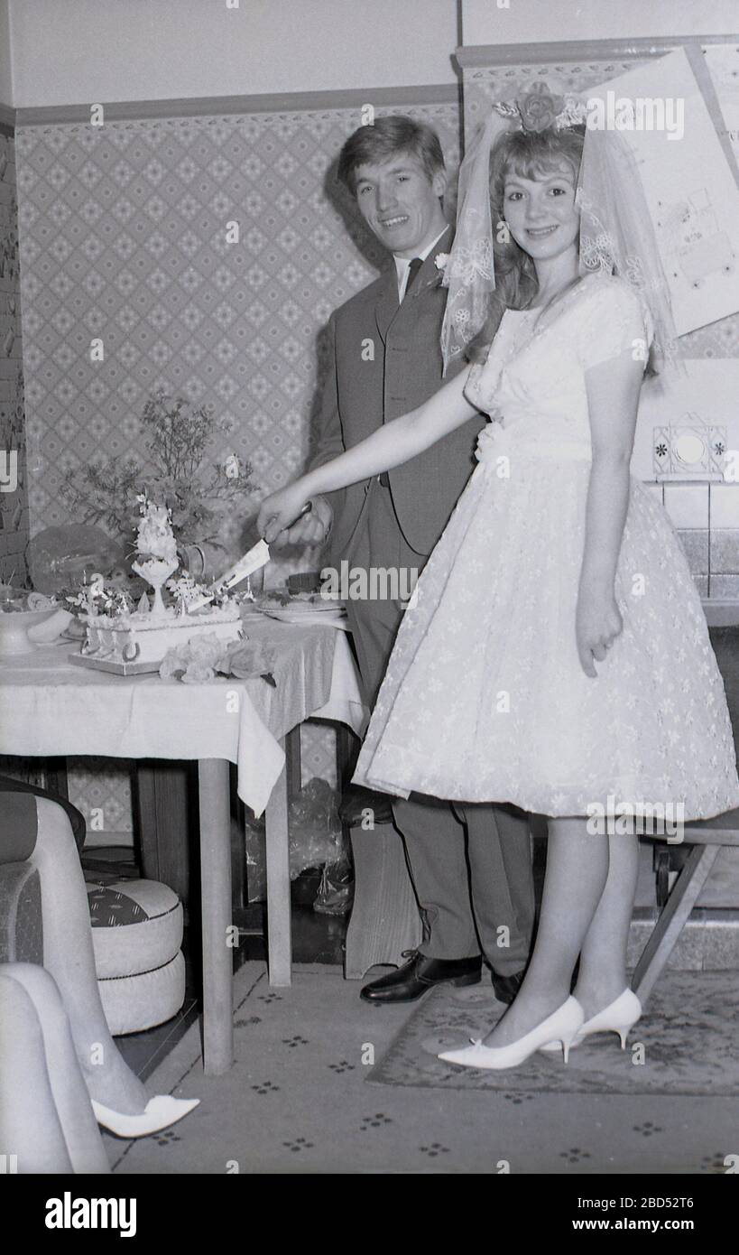 1960, historique, le grand jour, une adolescente mariée et son marié debout ensemble couper leur gâteau de mariage, Angleterre, Royaume-Uni. À cette époque comme dans les précédentes, les personnes se sont mariées à un âge plus jeune que les générations ultérieures. Banque D'Images