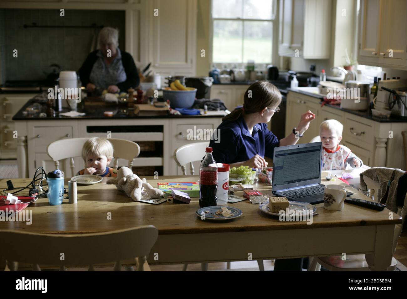 Mère nourrissant ses enfants dîner à la table de cuisine tout en travaillant à la maison pendant la période d'auto-isolation - 2020 COVID-19 pandémie de coronavirus Banque D'Images