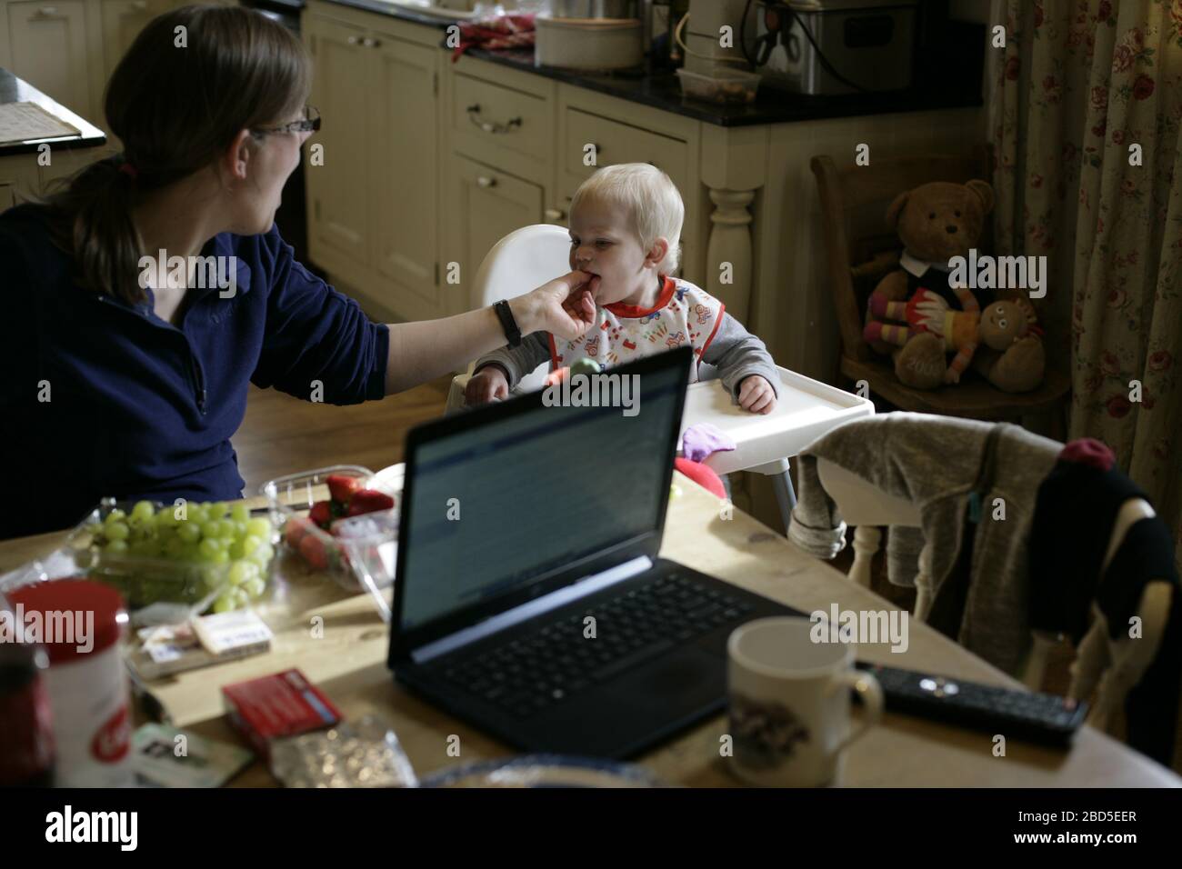 Mère nourrissant ses enfants dîner à la table de cuisine tout en travaillant à la maison pendant la période d'auto-isolation - 2020 COVID-19 pandémie de coronavirus Banque D'Images