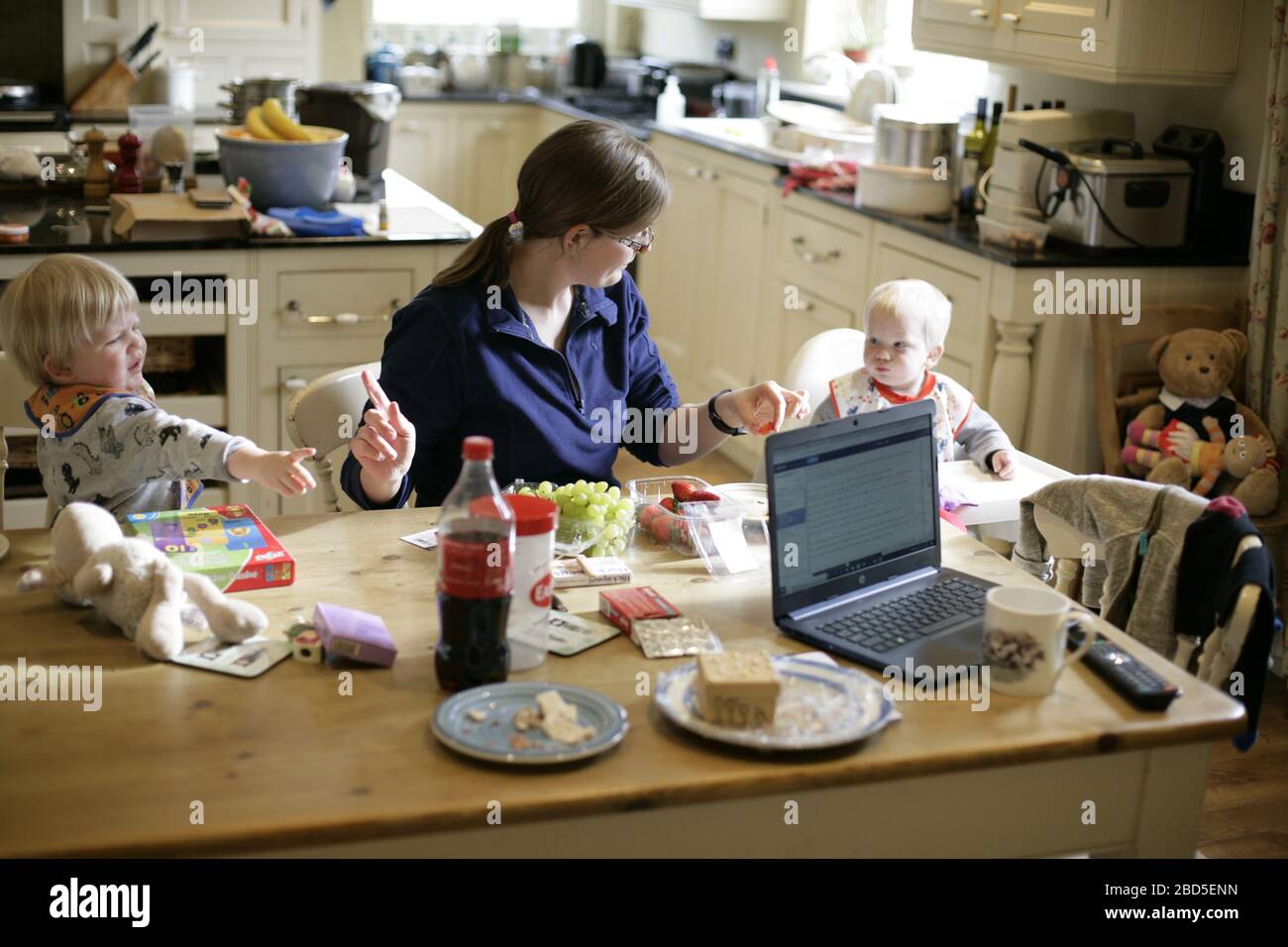 Mère nourrissant ses enfants dîner à la table de cuisine tout en travaillant à la maison pendant la période d'auto-isolation - 2020 COVID-19 pandémie de coronavirus Banque D'Images