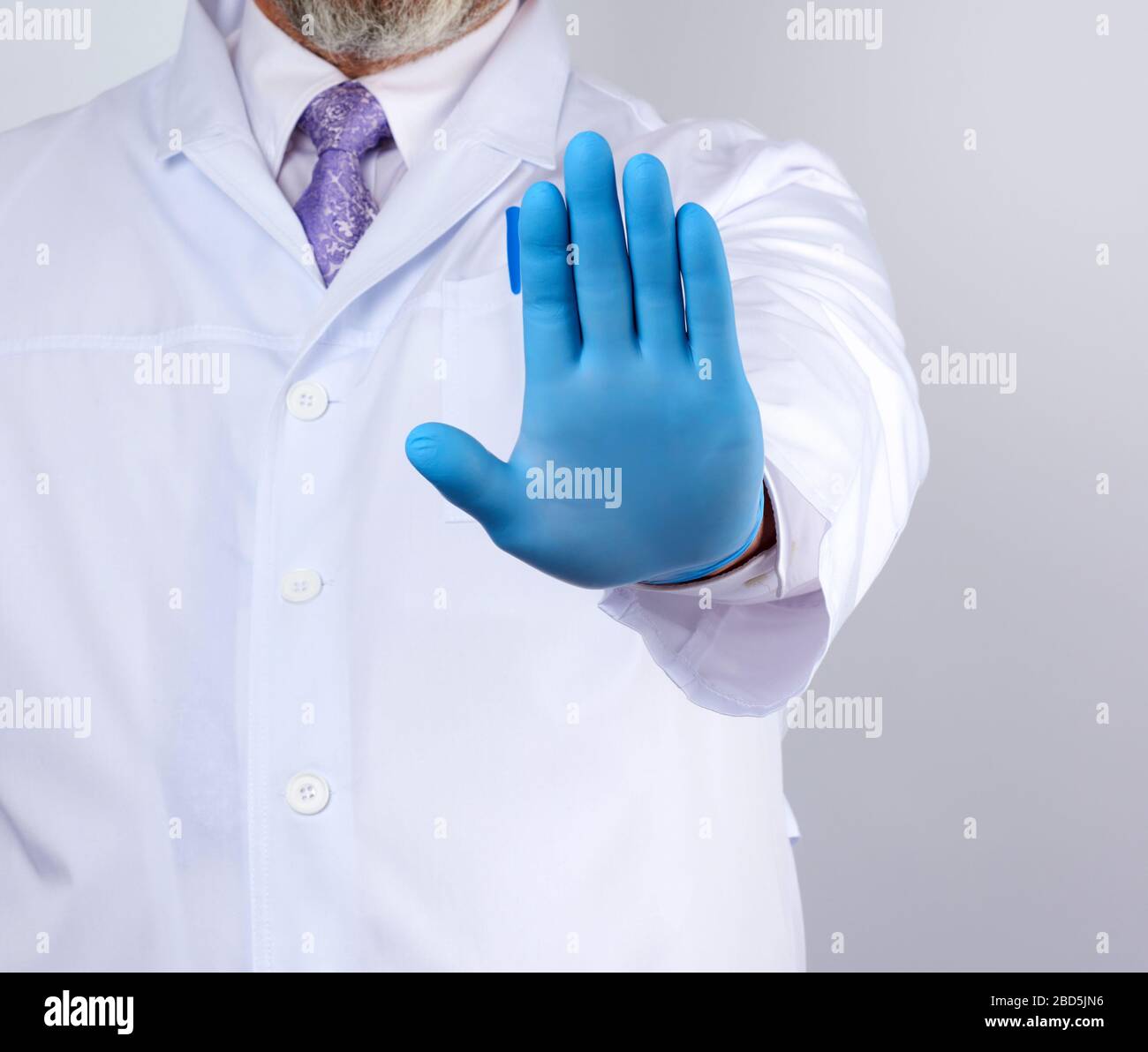 le médecin masculin dans un manteau blanc et des gants stériles bleus montre un geste d'arrêt avec sa main droite, fond blanc Banque D'Images