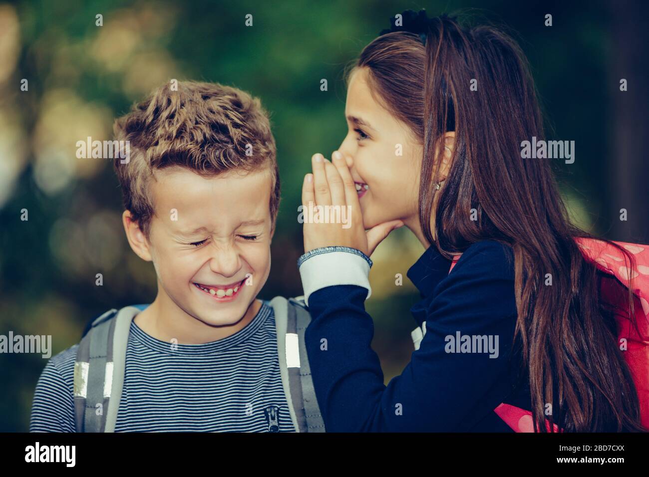 Fermez deux heureux amis avec des sacs à dos riant et s'amuser dans le parc. Une petite fille chuchote quelque chose dans une oreille de son ami avec le sourire an Banque D'Images