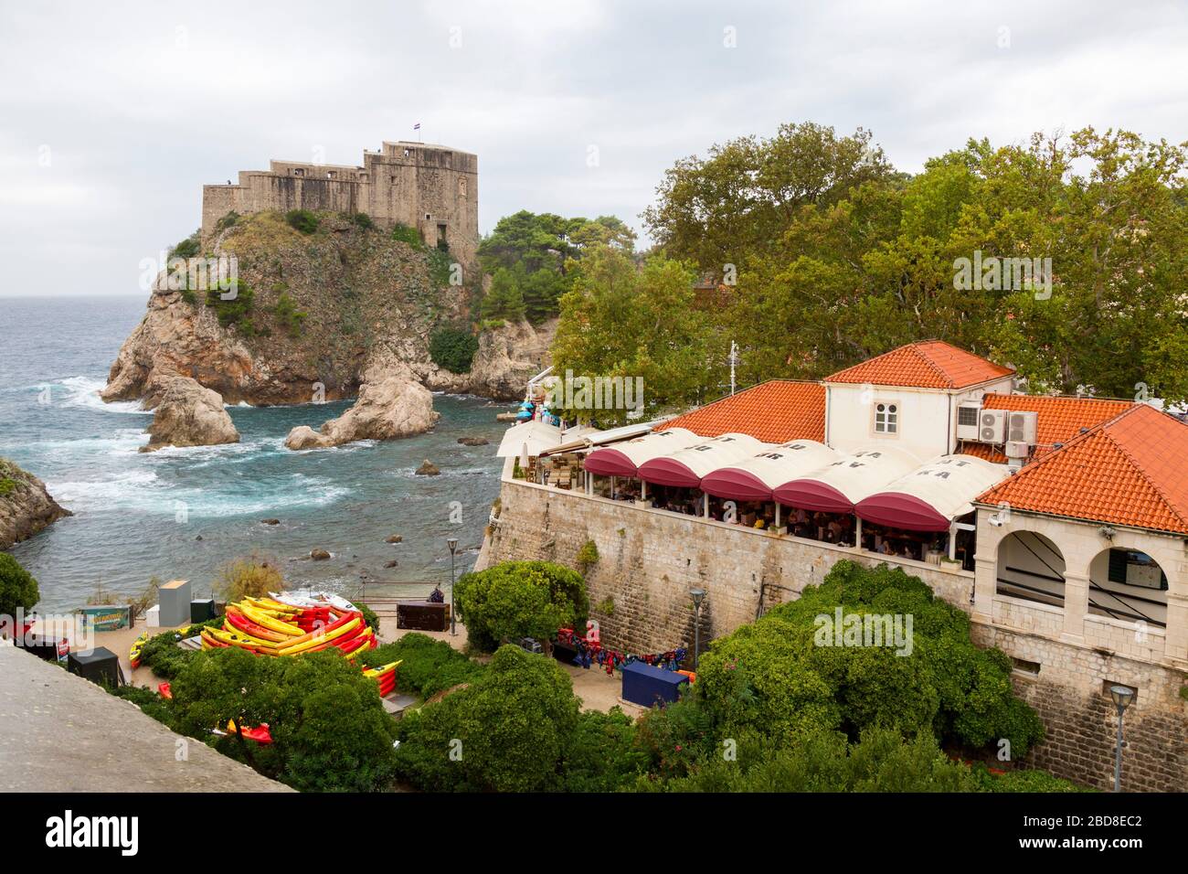Vue sur les murs historiques de la vieille ville de Dubrovnik, dans le port ouest. La vue comprend le fort Lovrijenac, la plage et le restaurant de bord de mer. Banque D'Images