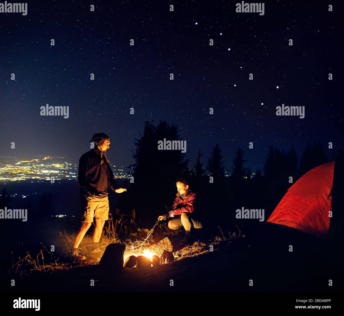 Couple heureux randonneurs ils chaud les mains près de camp sous ciel de nuit avec les étoiles et les lumières de la ville à l'arrière-plan. Banque D'Images