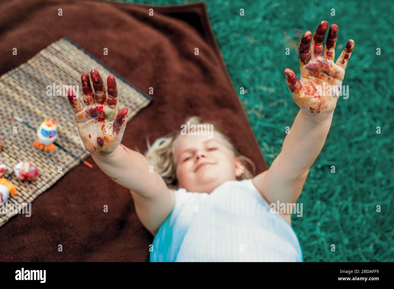 Mignonne petite fille layong sur la couverture sur l'herbe dans l'arrière-cour et montrer sa sale avec des mains peintes, peindre des œufs de Pâques. Banque D'Images