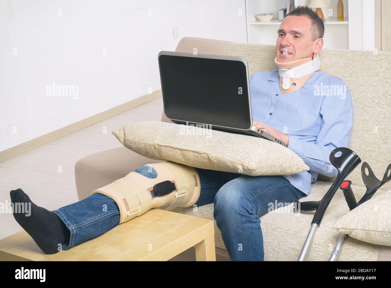 L'homme avec la jambe en neck brace, genou des cages et des béquilles de stabilisation et de soutien reposant sur un canapé avec ordinateur portable. Banque D'Images