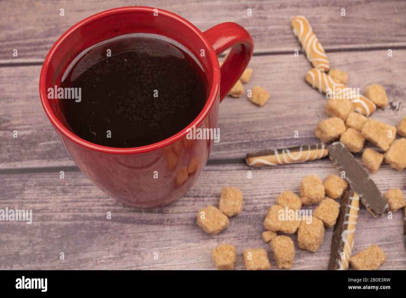 Tasse en céramique rouge avec thé, bâtonnets de biscuit avec chocolat et glaçage blanc, et morceaux de sucre de canne brun sur fond en bois. Gros plan Banque D'Images