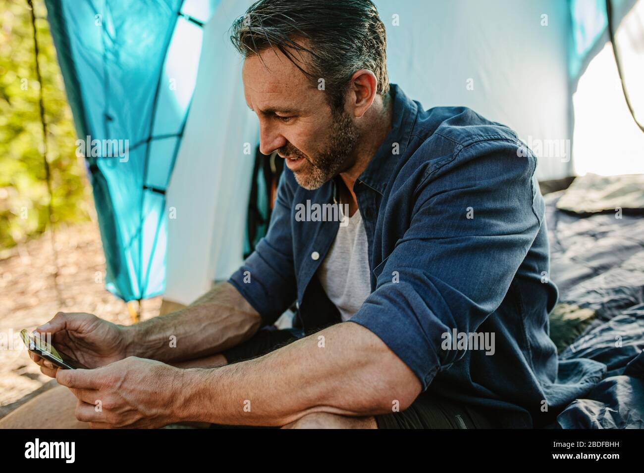 Homme mûr assis dans une tente et regardant son téléphone portable. Camping-car adulte moyen utilisant un smartphone au camping. Banque D'Images Homme mûr assis dans une tente et regardant son téléphone portable. Camping-car adulte moyen utilisant un smartphone au camping. Banque D'Images