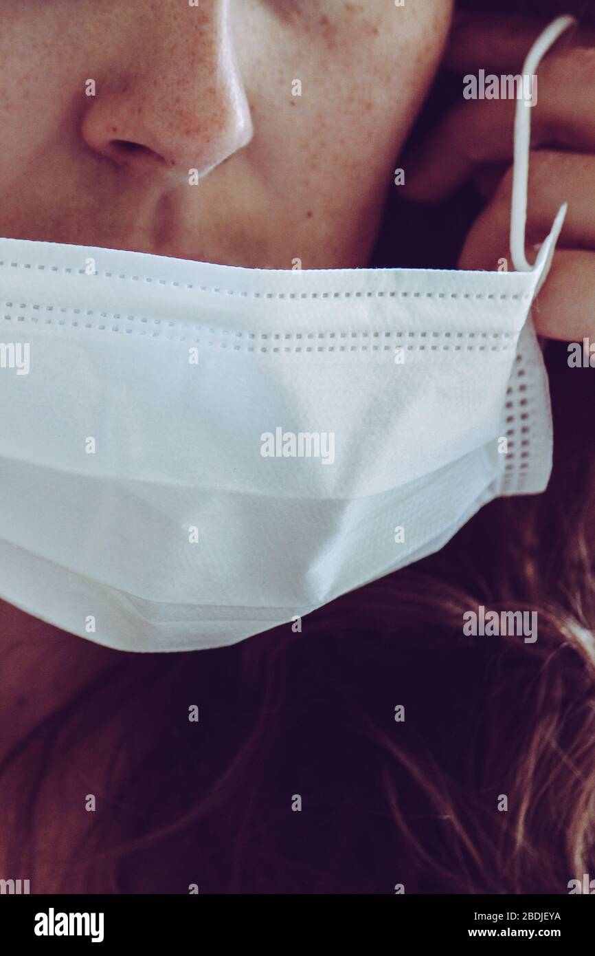Détail du visage de femme non reconnaissable portant un masque médical blanc. Partiellement flou, mise au point sélective, grain léger. Fille REDEHEAD avec des freckles. Coronavirus, COVID-19 quarantaine. Concept médecin/infirmière. Banque D'Images