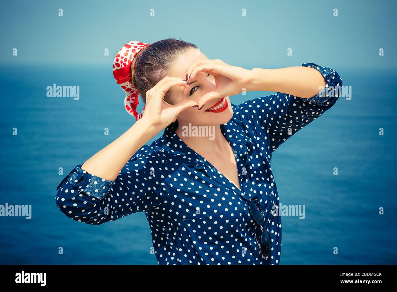 Amour. Gros plan portrait souriant jeune femme heureuse faisant signe de coeur, symbole avec des mains isolé paysage de mer arrière-plan. Expression positive de l'émotion humaine Banque D'Images