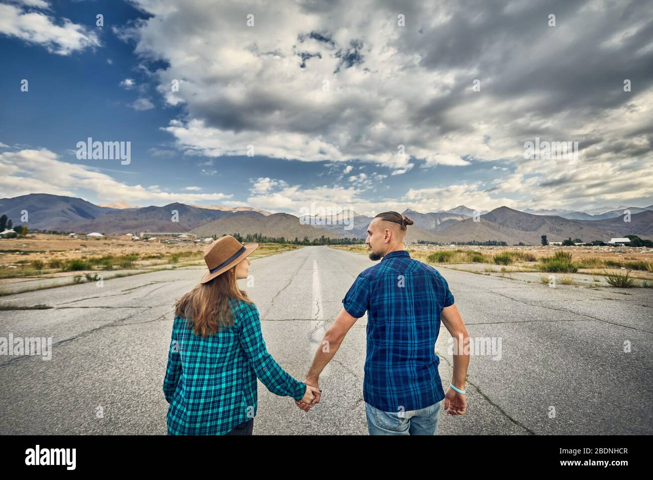 Heureux couple in checked shirt holding par les mains et marcher sur la large route asphaltée avec montagnes et fond de ciel nuageux Banque D'Images