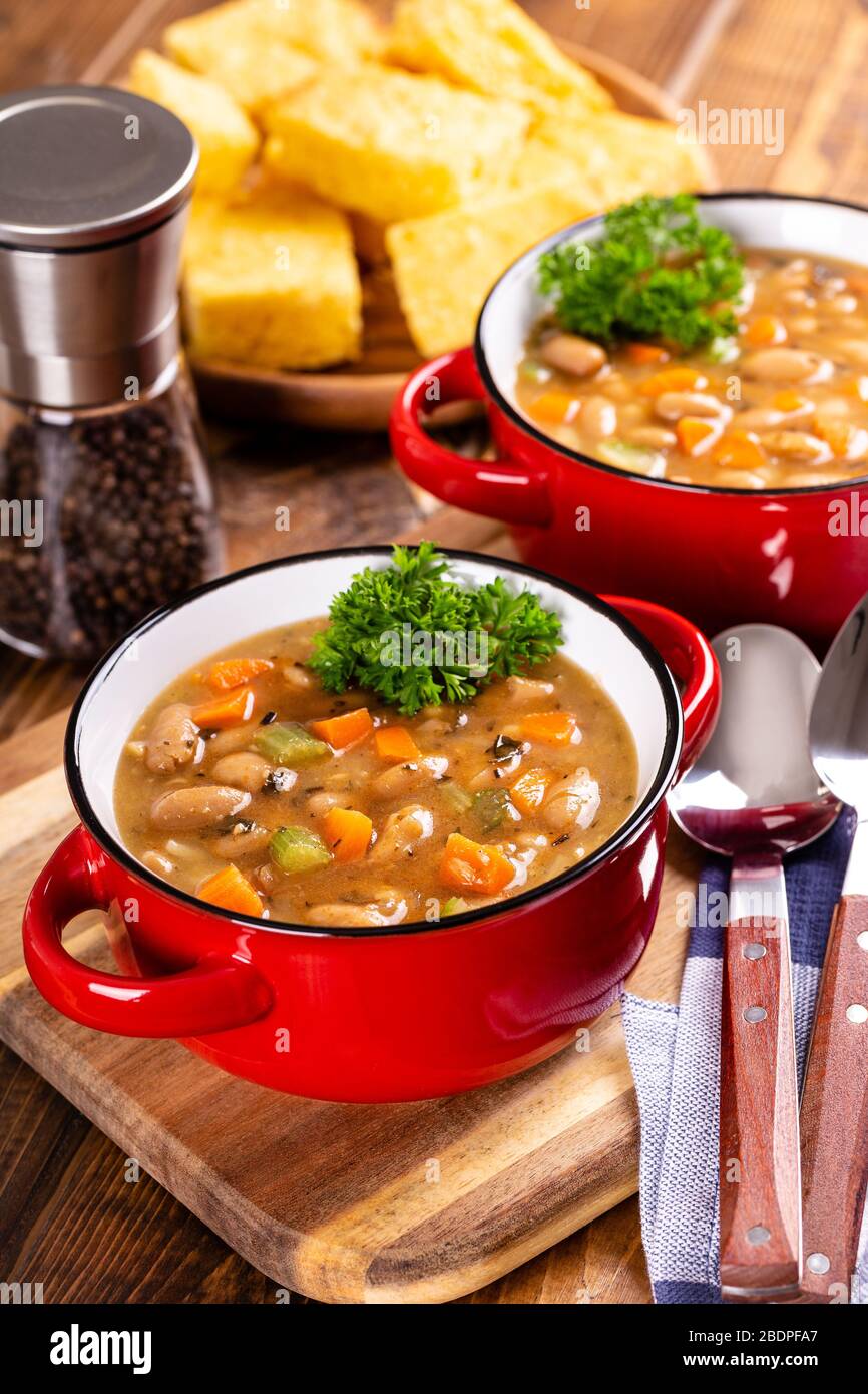 Soupe aux haricots et légumes savoureux dans un bol rouge avec pain de maïs en arrière-plan sur une table en bois Banque D'Images