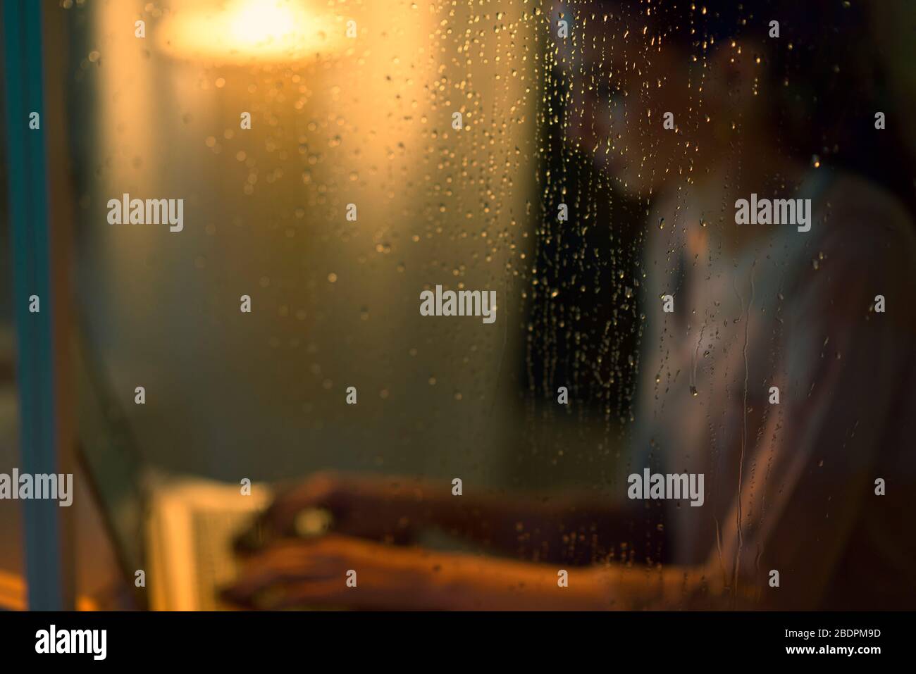 Jeune femme travaillant avec un ordinateur portable à côté d'une fenêtre sur une nuit de pluie, verre avec des gouttes au premier plan Banque D'Images