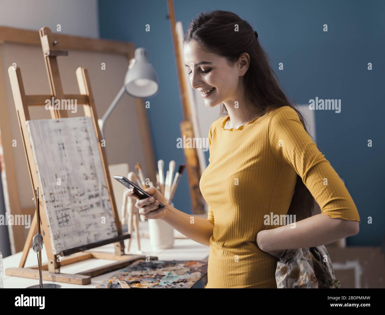 Heureuse jeune artiste dans son studio d'art, elle se tient à côté de sa peinture et discute avec son smartphone Banque D'Images