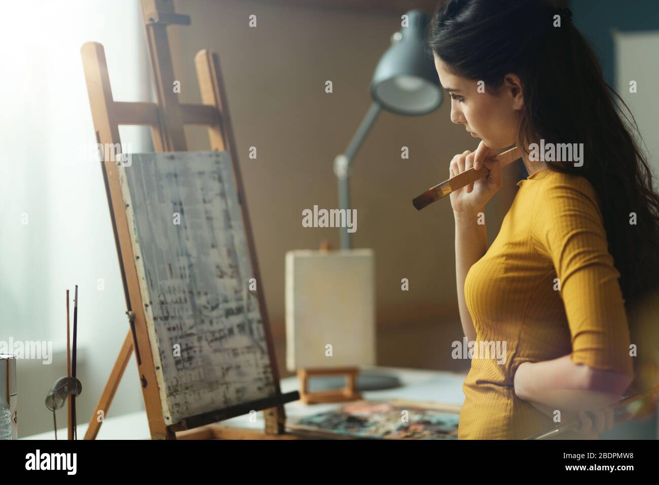 Jeune artiste féminine travaillant dans le studio d'art et en vérifiant sa peinture, elle tient une brosse et une pensée Banque D'Images