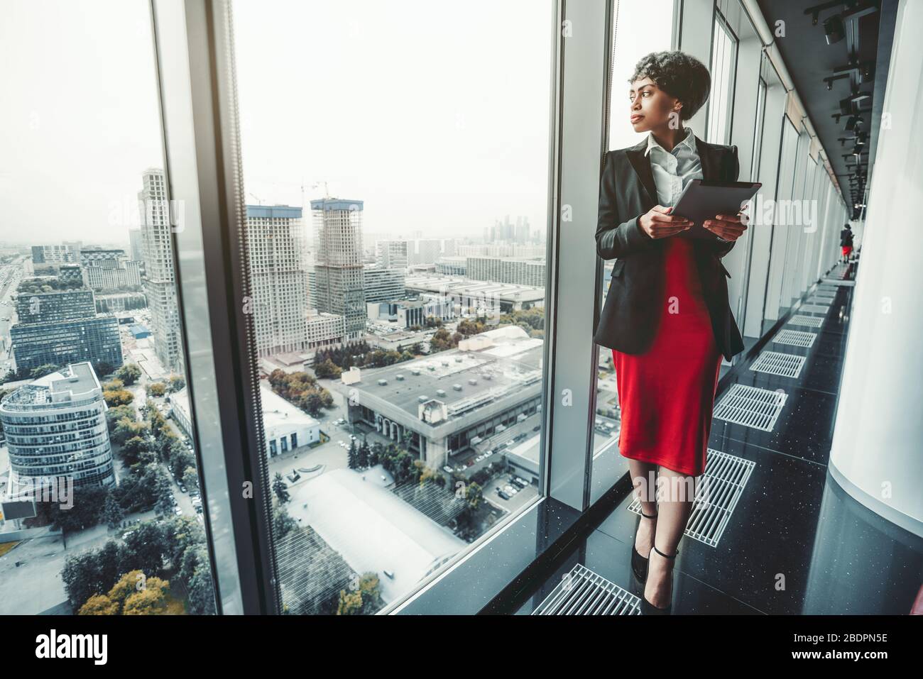 Une femme d'affaires afro-américaine de bonne qualité dans une jupe rouge et une veste noire utilise une tablette numérique tout en se penchant contre une fenêtre panoramique Banque D'Images