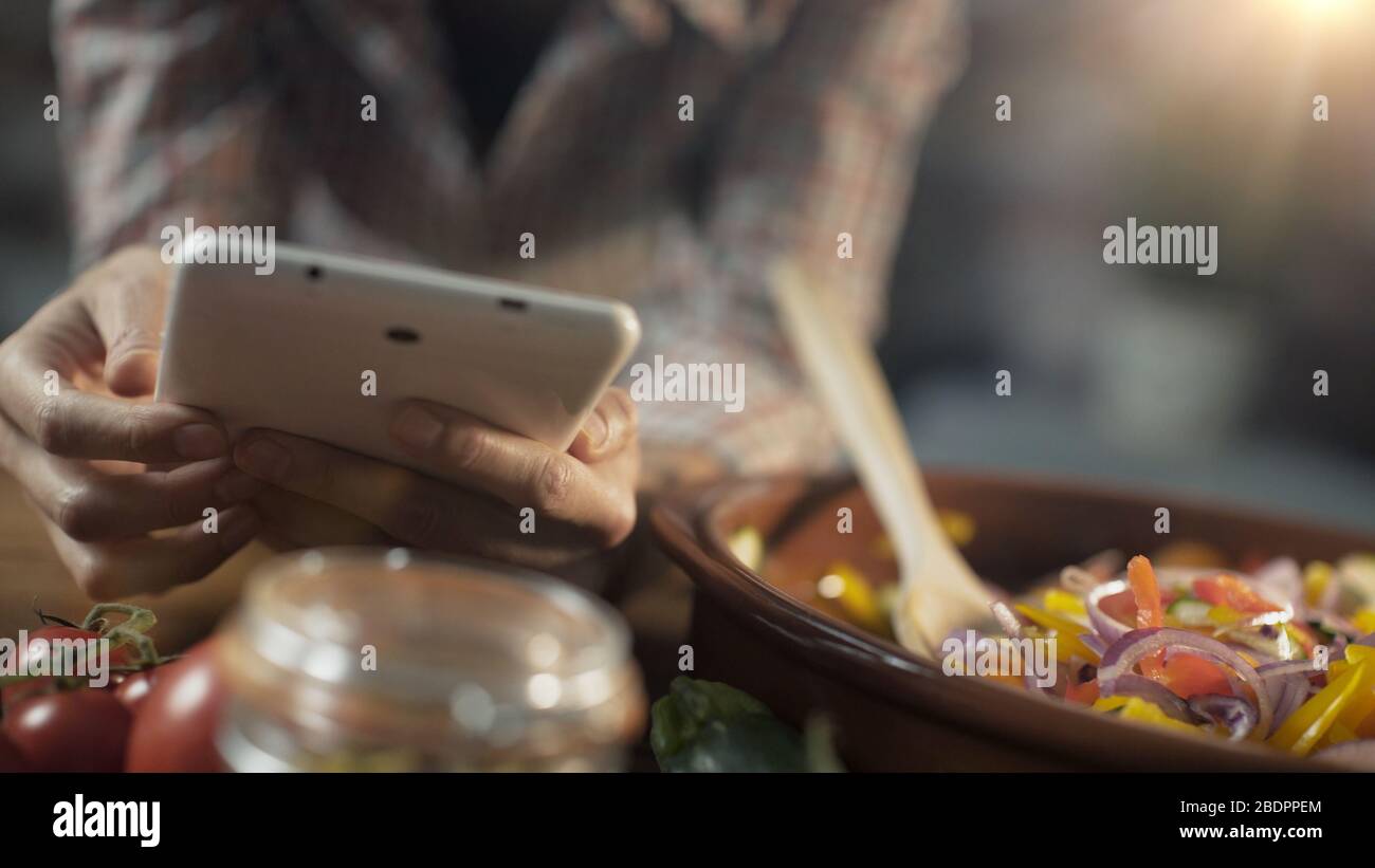 Femme recherchant des recettes saines en ligne sur sa tablette numérique et la cuisine de légumes frais mélangés dans une casserole d'argile Banque D'Images