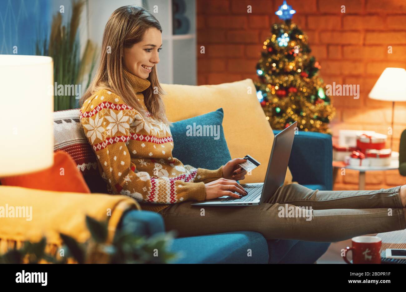 Heureuse jeune femme assise sur le canapé à la maison et faisant des achats en ligne pour des cadeaux de Noël, elle se connecte avec son ordinateur portable et détient un crédit c Banque D'Images