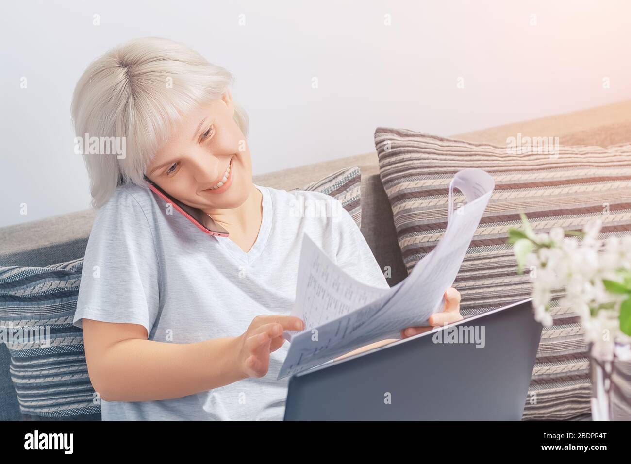 Une jolie fille souriante avec des cheveux blonds tient des documents financiers d'une part, les retourne avec l'autre main tout en parlant sur le téléphone dans la maison Banque D'Images