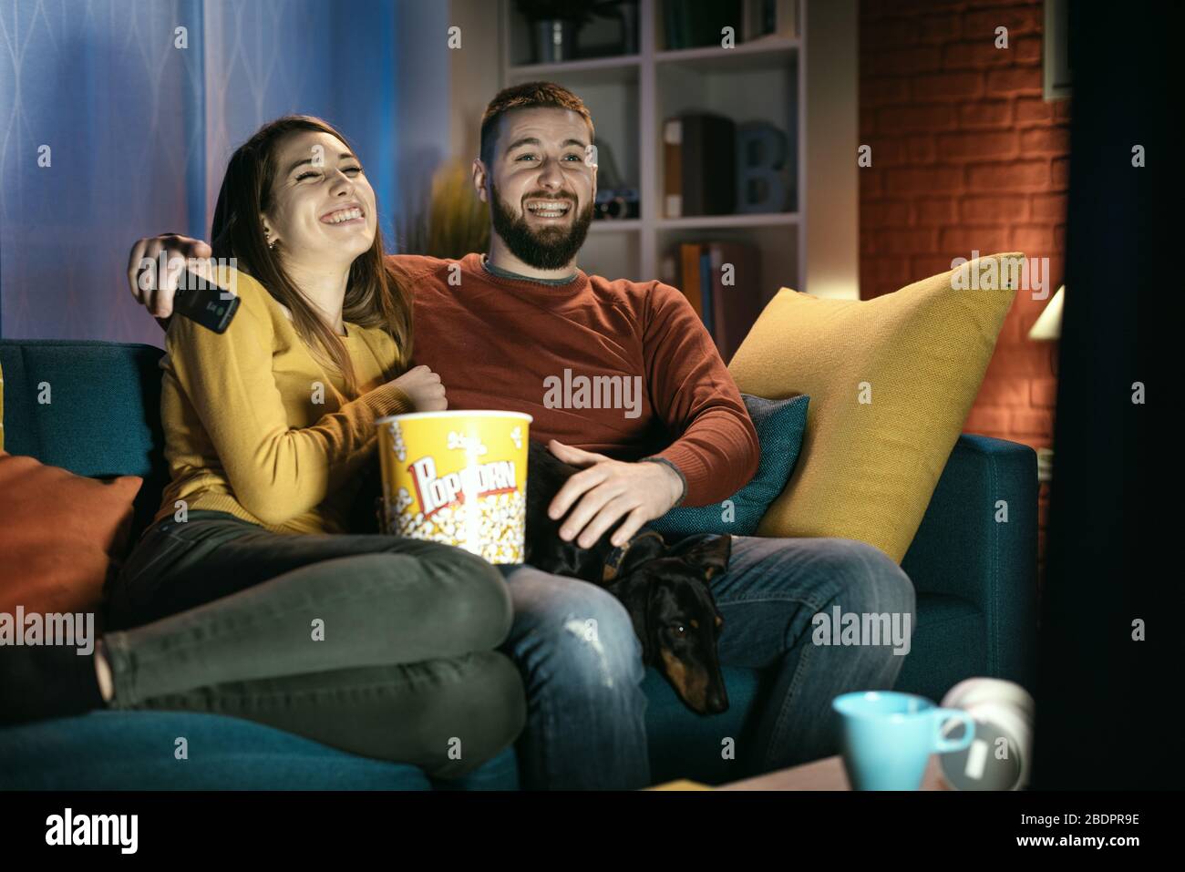 Heureux couple se reposant sur le canapé à la maison avec leur chien: Ils regardent des émissions de télévision, manger du pop-corn et rire ensemble Banque D'Images