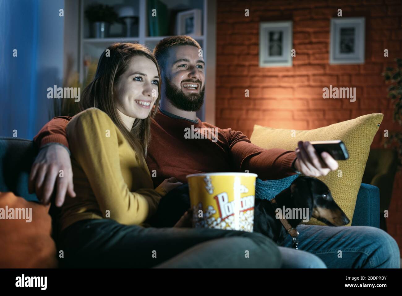 Heureux couple se reposant sur le canapé à la maison avec leur chien, ils regardent des émissions de télévision et mangent du pop-corn, il tient la télécommande Banque D'Images